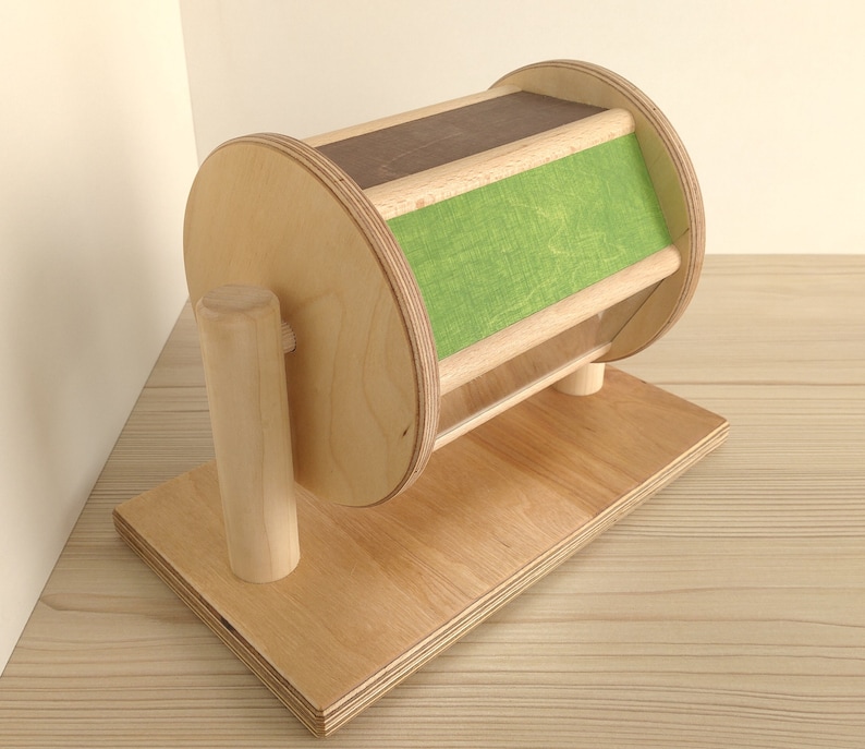 May include: A wooden toy with a spinning wheel and a green fabric panel. The wheel is mounted on a wooden base with a wooden post.