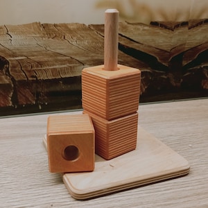 May include: A wooden stacking toy with three square blocks and a wooden dowel. The blocks have a light brown color and a grooved texture. The toy is on a wooden base.