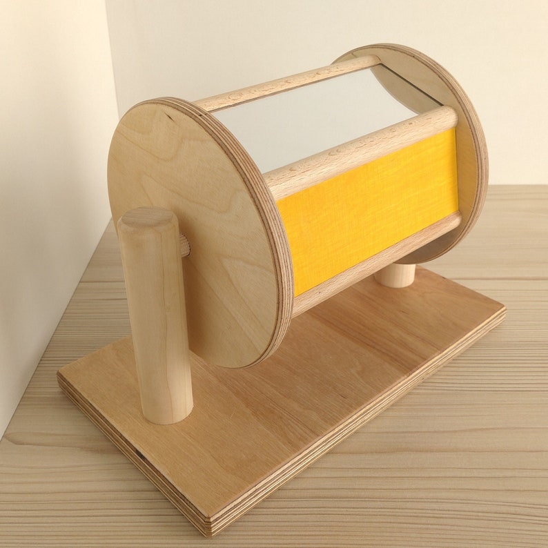 May include: Wooden toy with a mirror and yellow panel, mounted on a wooden base. The toy is designed to spin and reflect light.