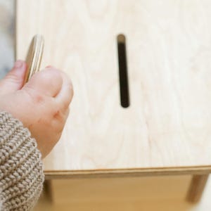 Könnte beinhalten: Ein helles Holz-Sparschwein mit einem Schlitz oben. Eine Hand, die eine Holz-Münze hält, ist dabei, sie in den Schlitz zu stecken. Die Bank hat ein schlichtes, minimalistisches Design, mit quadratischer Form und vier Beinen.