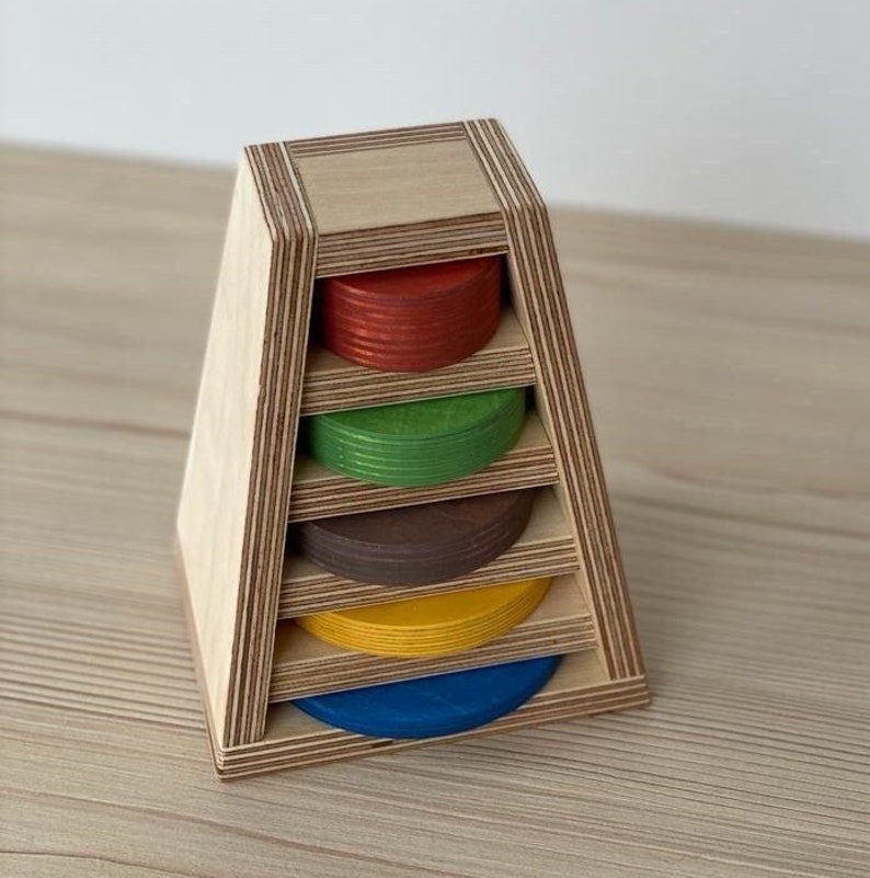 May include: Wooden stacking toy with five colorful rings. The rings are red, green, brown, yellow, and blue. The toy is made of plywood and has a triangular base.