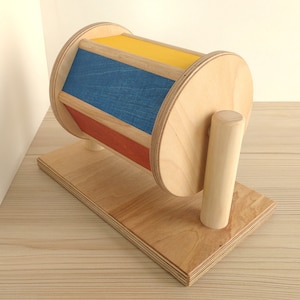 May include: A wooden toy with a spinning drum. The drum is painted with red, blue, and yellow stripes. The toy is on a wooden base with a wooden handle.