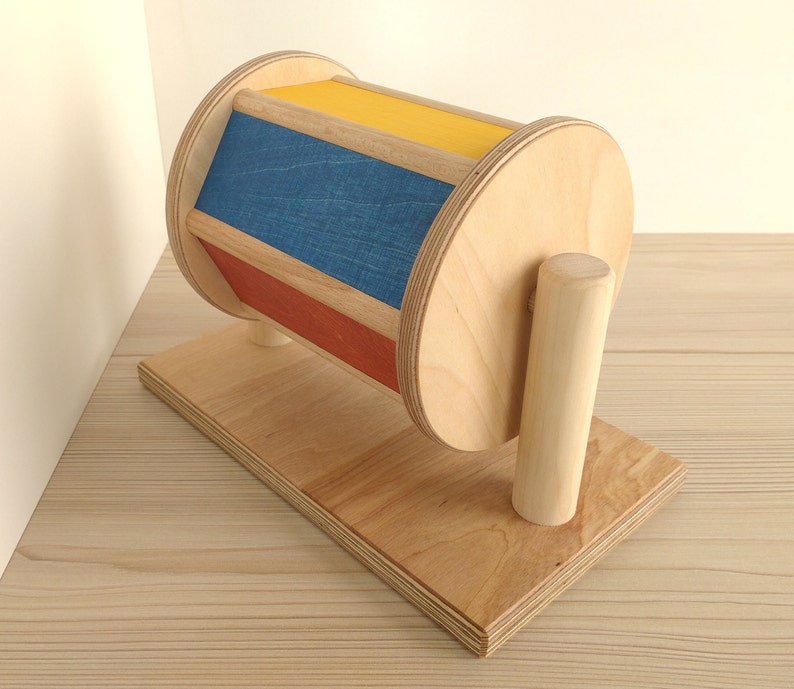 May include: A wooden toy with a spinning drum. The drum is painted with red, blue, and yellow stripes. The toy is on a wooden base with a wooden handle.