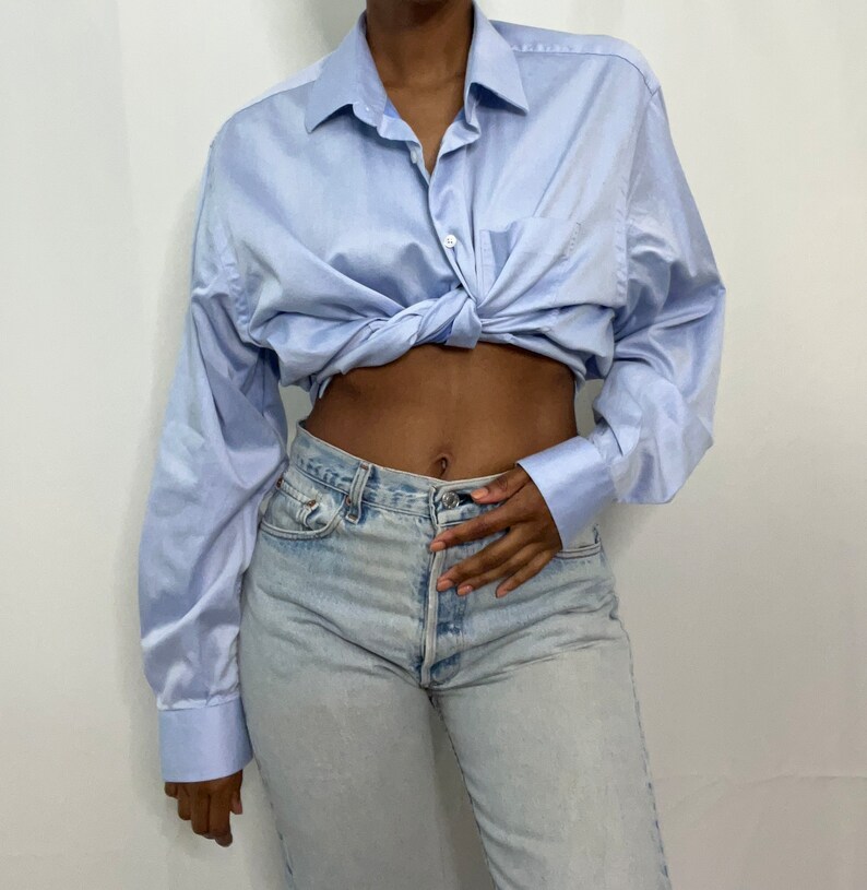 May include: A light blue long-sleeved button-down shirt tied at the waist over light blue denim jeans.
