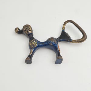 Puede incluir: Un abridor de botellas de metal azul y dorado con forma de perro estilizado. El abridor tiene un diseño distintivo con una cabeza y una cola texturizadas en relieve.