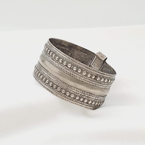 Puede incluir: Un brazalete ancho de color plateado. El brazalete presenta una serie de bandas decorativas con patrones de cuentas y texturas. Un cierre rectangular asegura el brazalete. La joya se exhibe sobre un fondo blanco.