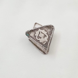 Puede incluir: Colgante triangular plateado con un diseño floral ornamentado. El colgante tiene un cierre de bisagra y un borde detallado. El metal tiene un aspecto ligeramente envejecido, lo que sugiere que es vintage o antiguo.