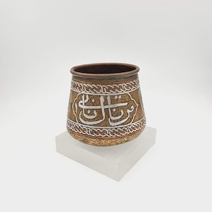 May include: A copper-toned metal cup featuring intricate embossed patterns and silver-colored Arabic calligraphy centered on the exterior. The vessel has a wide, rounded body with a dark interior rim and a decorative braided border design.