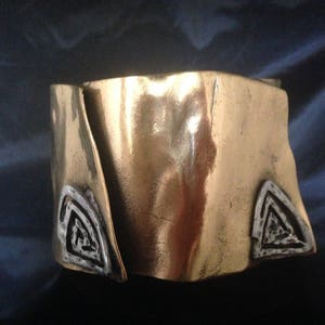 Pode incluir: Um bracelete de metal dourado com textura martelada e dois desenhos geométricos prateados.