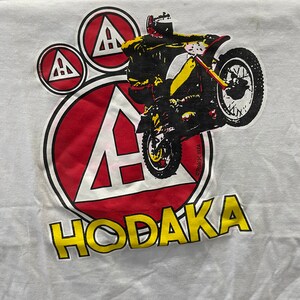 May include: White t-shirt with a red, white, and yellow graphic of a motorcycle with the text "HODAKA" and "©ROACH 1974".