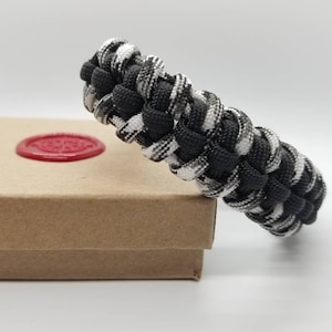 May include: Black and white camouflage paracord bracelet. The bracelet is made with a woven pattern and is a popular style for outdoor enthusiasts.