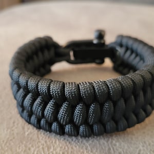 May include: A black paracord bracelet with a black metal clasp. The bracelet is made of woven paracord and has a simple design.