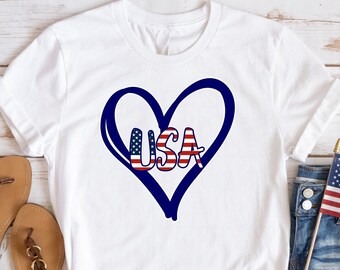 May include: White t-shirt with a blue heart design and the word "USA" in red, white, and blue. The shirt is paired with denim shorts, sandals, and a small American flag. The design is patriotic and suitable for Independence Day.