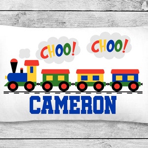 May include: A white pillow with a colorful train design and the name "CAMERON" printed in blue. The train has a blue engine, yellow caboose, green and yellow cars, and red wheels. The words "CHOO!" are printed in red, yellow, and blue on white clouds.