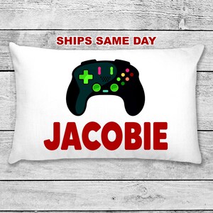 May include: White pillow with a black and green video game controller graphic and the name "Jacobie" in red letters.