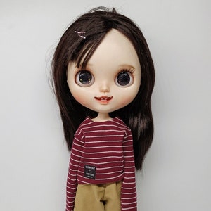 May include: A doll with long brown hair wearing a red and white striped long-sleeved shirt, beige corduroy pants, and black sneakers. The shirt has a small black patch with the text "MOMENT IS EVERYTHING".