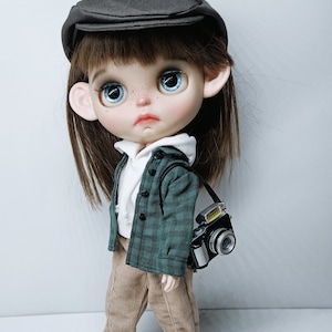 May include: A doll wearing a green and black plaid shirt, brown pants, and black and white sneakers. The doll is also wearing a black cap and has brown hair. The doll is holding a camera.