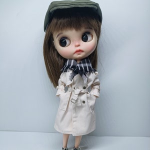 May include: A doll wearing a white trench coat, a black and white checked scarf, a green newsboy cap, and black trainers.