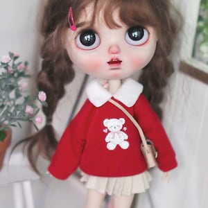 Può includere: Un maglione rosso per bambola con un orsacchiotto bianco ricamato sul davanti. Il maglione ha un colletto bianco e un bordo bianco intorno alle maniche. La bambola indossa una gonna beige e ha i capelli castani.