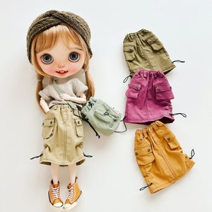 Könnte beinhalten: Eine Puppe trägt einen braunen und grünen Rock mit einem braunen Stirnband und einem grünen Rucksack. Die Puppe steht auf einer weißen Oberfläche. Es gibt drei weitere Röcke auf dem Bild: einen grünen, einen burgunderfarbenen und einen orangen.