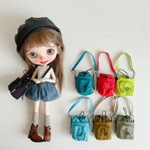 Può includere: Una bambola con capelli neri e un berretto nero tiene uno zainetto nero di piccole dimensioni. Sei piccole borse a cordoncino di diversi colori sono disposte in fila su una superficie bianca. Le borse hanno spalline regolabili e sono realizzate in un materiale simile alla tela.