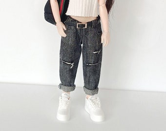 May include: A doll with brown hair styled in two buns, wearing a white turtleneck sweater, blue jeans with rips, a black bag with a red and green strap, and white sneakers.