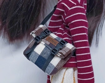 May include: A doll wearing a red and white striped long-sleeved shirt, white pants with gold stripes, and black shoes. The doll is carrying a black and white plaid bag with black buckles and the word "XING" printed on it.
