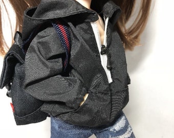 May include: A black hooded jacket with a red and blue stripe on the zipper. The jacket is worn by a doll with blue jeans and a black backpack.