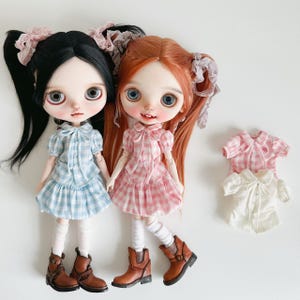 May include: Two dolls with large eyes and pigtails, one with black hair and the other with red hair, wearing matching plaid dresses and brown boots. Additional doll outfits in pink and white are also present.