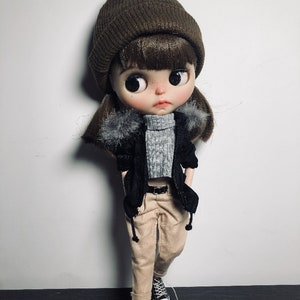 May include: A brown beanie with a black jacket and beige trousers. The doll is wearing black and white trainers.