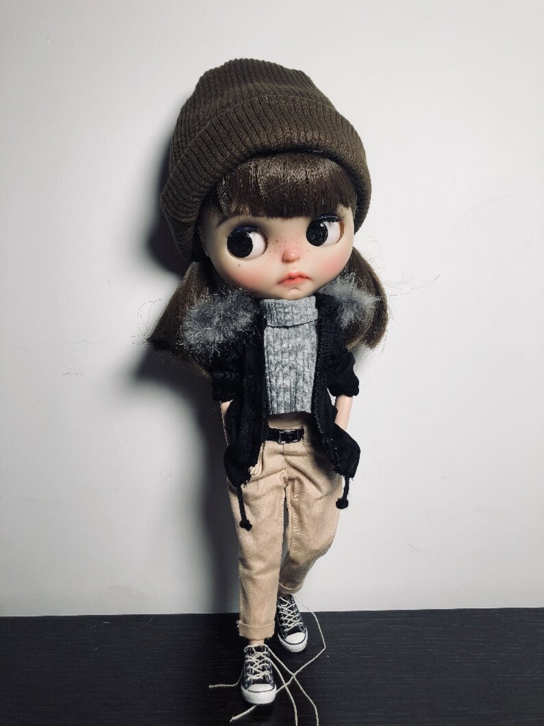 May include: A brown beanie with a black jacket and beige trousers. The doll is wearing black and white trainers.