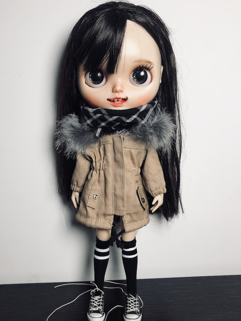 May include: A doll with long black hair wearing a brown jacket with a fur trim, a black and white checked scarf, black and white striped socks, and black trainers.