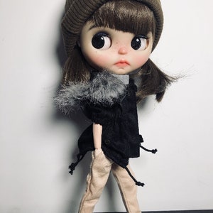 May include: A doll wearing a brown beanie, a black jacket with a faux fur trim, beige trousers, and black and white trainers.