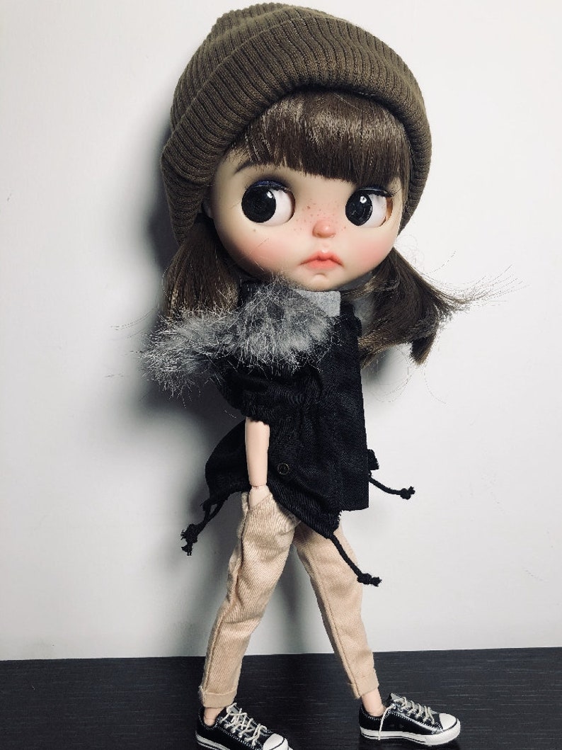 May include: A doll wearing a brown beanie, a black jacket with a faux fur trim, beige trousers, and black and white trainers.