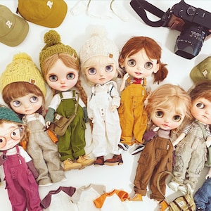 Op de afbeelding: Zeven poppen met grote ogen en bruin haar, die overalls dragen en verschillende accessoires bij zich hebben. De poppen zijn op een wit oppervlak gerangschikt met een camera en hoeden op de achtergrond.