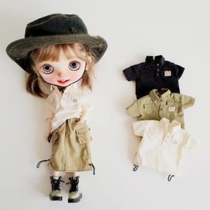 Könnte beinhalten: Eine Puppe trägt ein weißes Hemd, einen Khakirock und einen grünen Hut. Vor der Puppe liegen drei weitere Hemden: ein schwarzes, ein khakifarbenes und ein weißes.