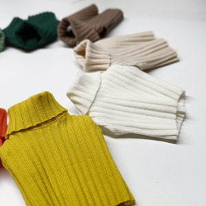 May include: A collection of ten different coloured ribbed knit turtleneck sweaters. The sweaters are arranged in a circle on a white background.
