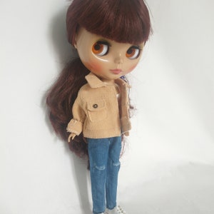 May include: A doll with long brown hair wearing a light brown corduroy jacket, blue jeans with a distressed detail, and white shoes.