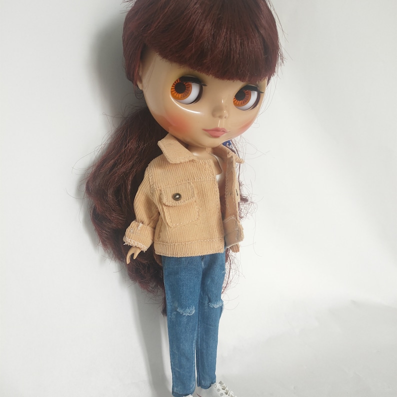 May include: A doll with long brown hair wearing a light brown corduroy jacket, blue jeans with a distressed detail, and white shoes.