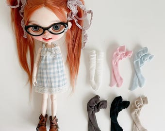 Può includere: Una bambola con capelli rossi a codini, con occhiali e un vestito a quadri blu e bianco. Vengono esposti diversi paia di calze per bambole bianche, rosa, blu, grigie, nere e beige.