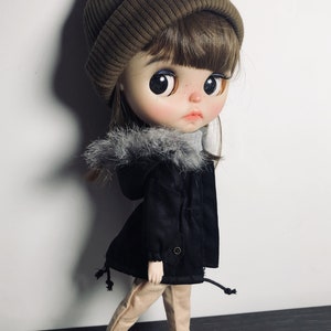 May include: A doll wearing a brown beanie, a black jacket with a faux fur trim, beige trousers, and black trainers.