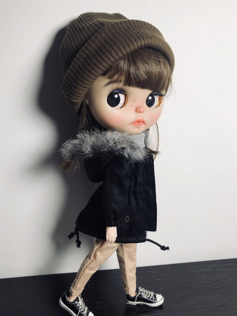 May include: A doll wearing a brown beanie, a black jacket with a faux fur trim, beige trousers, and black trainers.