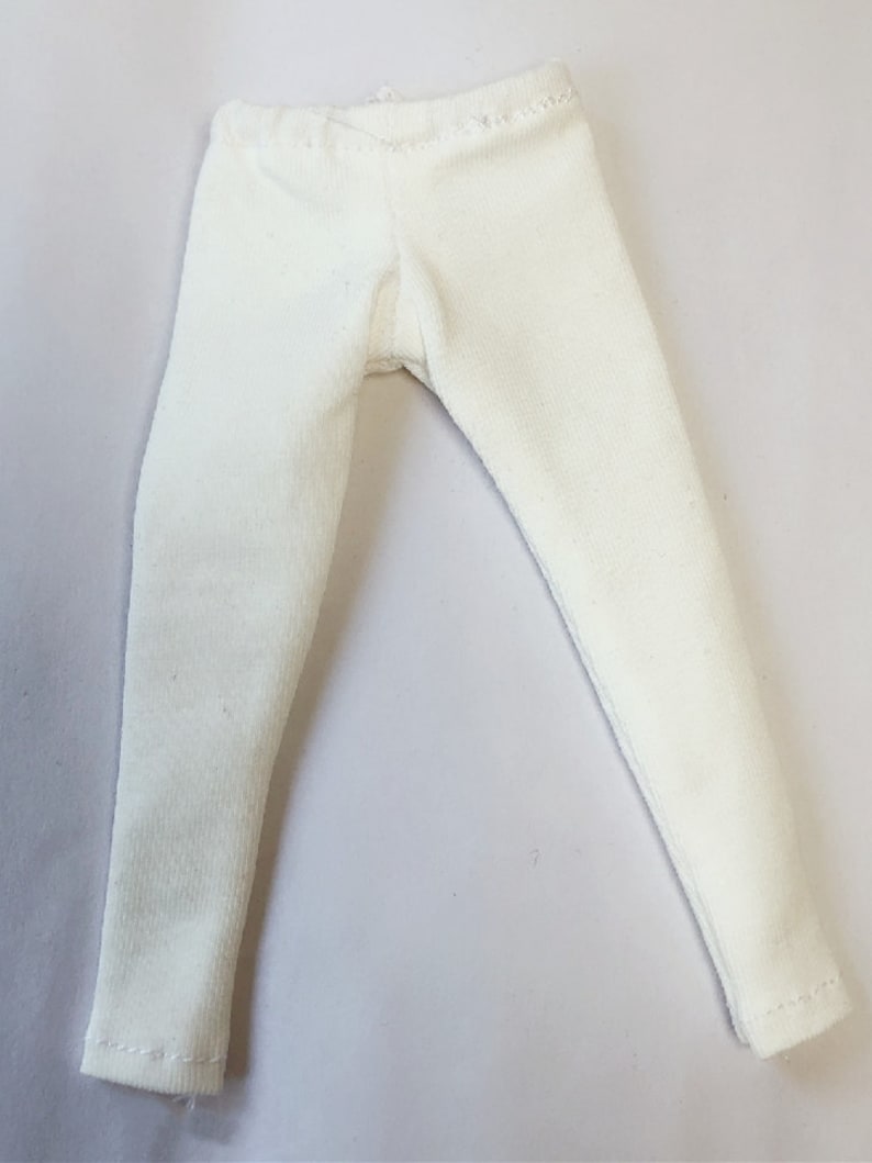Może przedstawiać: Białe legginsy dla lalki wykonane z miękkiej, elastycznej tkaniny. Legginsy są gładkie i mają prosty design.