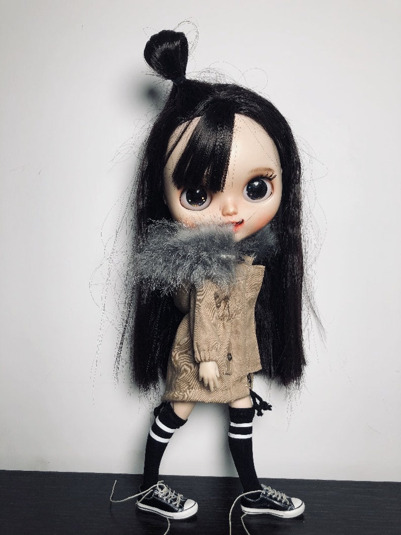 May include: A doll with long black hair, wearing a brown jacket with a grey fur collar, black and white striped socks, and black trainers.