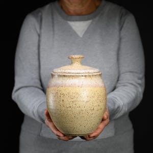 May include: A ceramic jar with a lid, featuring a speckled beige and yellow body that transitions to a soft pinkish-white glaze near the rim. The vessel has a rounded, bulbous shape and a small, knob-like handle on the center of the lid.