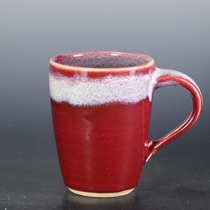 May include: A handmade ceramic mug with a deep red glaze. The top edge features a white band, and the handle is the same red color. The mug is approximately 4 inches tall and suitable for hot or cold beverages.