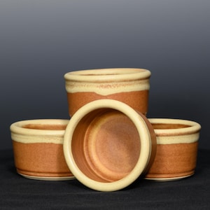 May include: Four ceramic ramekins with a rustic design. The ramekins are a warm brown color with a cream-colored band around the top rim. The interior of the ramekins is also brown. These small bowls are ideal for baking and serving individual portions.