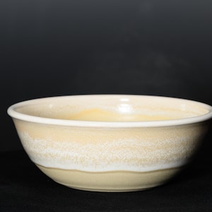 May include: A ceramic bowl with a cream-colored exterior and a white rim. The bowl has a textured surface with a band of white near the base. The interior of the bowl is a light yellow color. The bowl is set against a dark background.