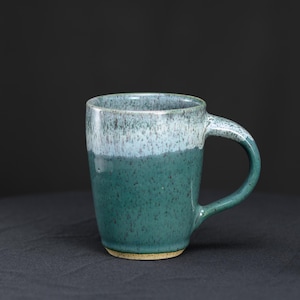 May include: A ceramic mug featuring a two-tone glaze with a dark teal base and a lighter, speckled white upper section. The mug has a curved handle on the right side and a smooth, cylindrical body with a visible unglazed rim at the base.