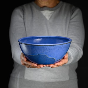 May include: A round ceramic bowl with a smooth, deep blue glaze. The bowl features a slightly darker blue tone near the base, creating a subtle gradient effect. The rim is defined by a thin, dark, and textured edge that contrasts with the glossy finish.